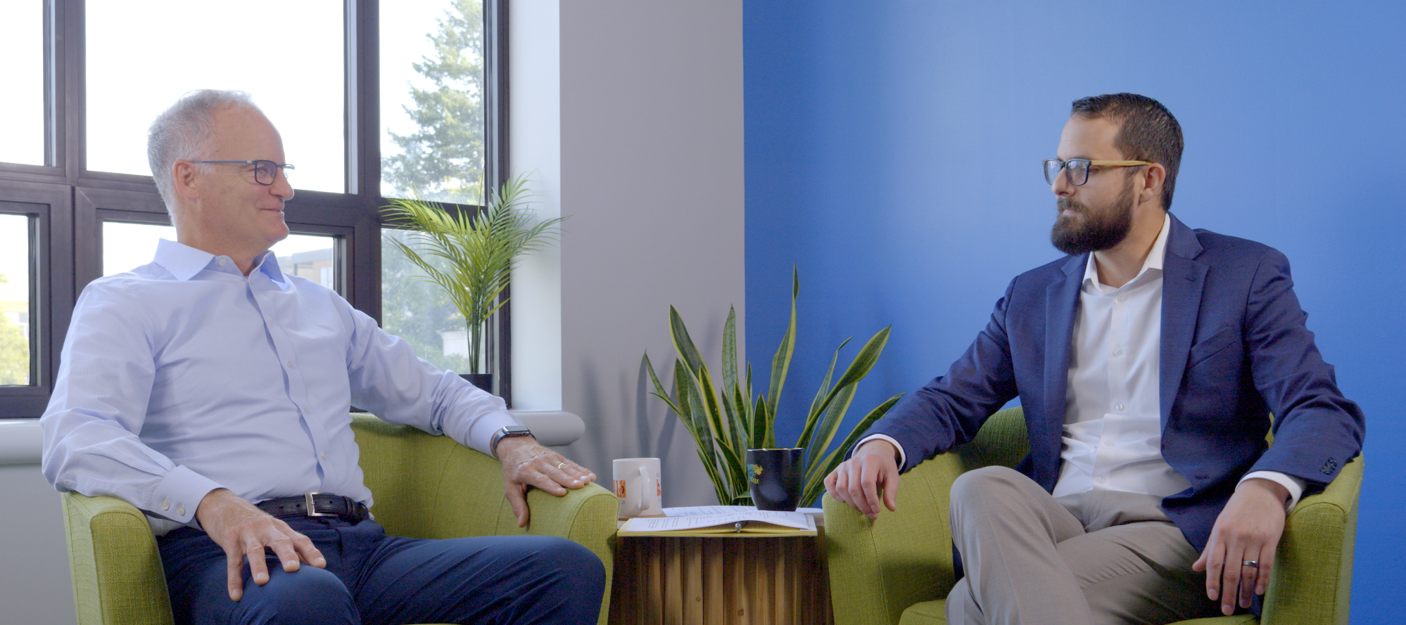 Two men sit in chairs facing each other at a wide angle, chatting over the coffee cups between them